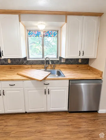 White cabinetry with black hardware, wood-finish countertops, stainless steel sink, stainless steel dishwasher, and wood-finish flooring