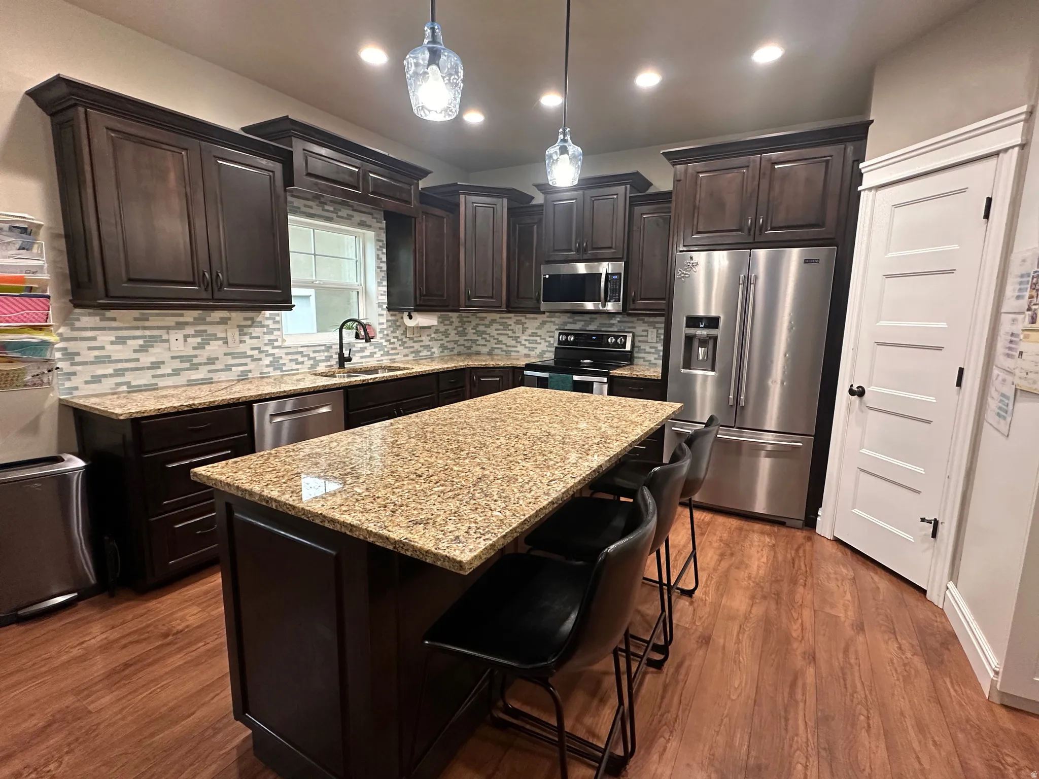 Kitchen with dark wood finish cabinetry, stainless steel appliances, a kitchen island, light stone counters, and decorative light fixtures