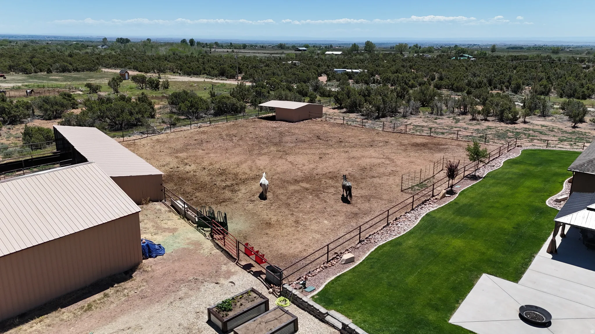 Expansive fenced pasture area featuring two detached outbuildings with metal roofs, a separate covered shelter, and a manicured lawn with a concrete patio