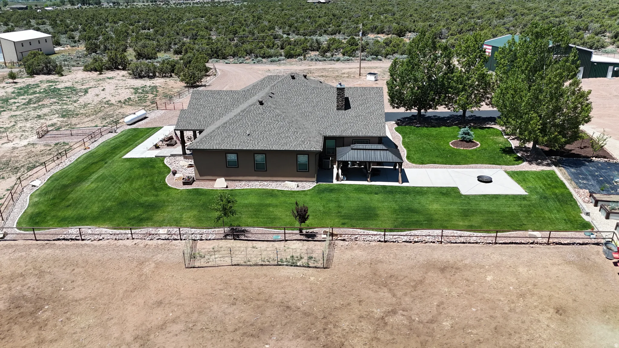 Expansive green lawn bordering the property, featuring a covered patio with a dark-framed roof structure, a concrete patio area, and mature trees