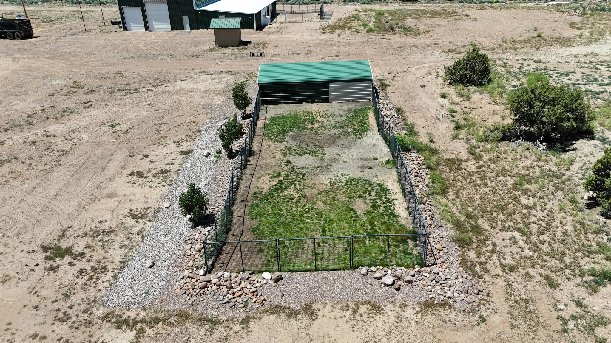 Fenced enclosure featuring a green-roofed shelter, partial vegetation, and surrounding gravel and rock landscaping