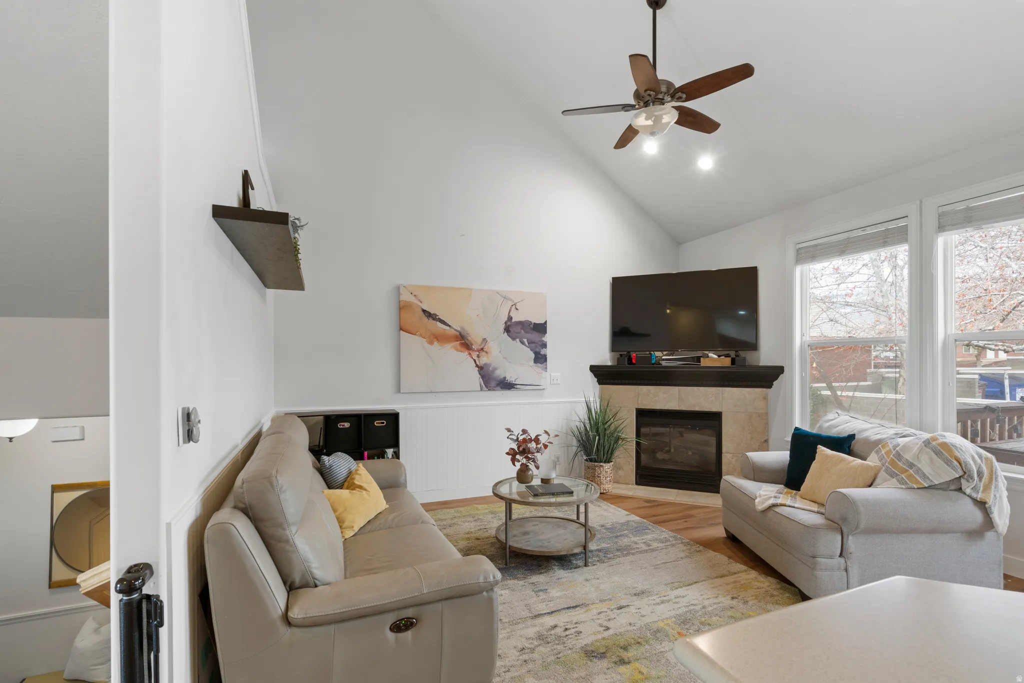 Living room featuring a fireplace, wood finished floors, ceiling fan, wainscoting, and lofted ceiling