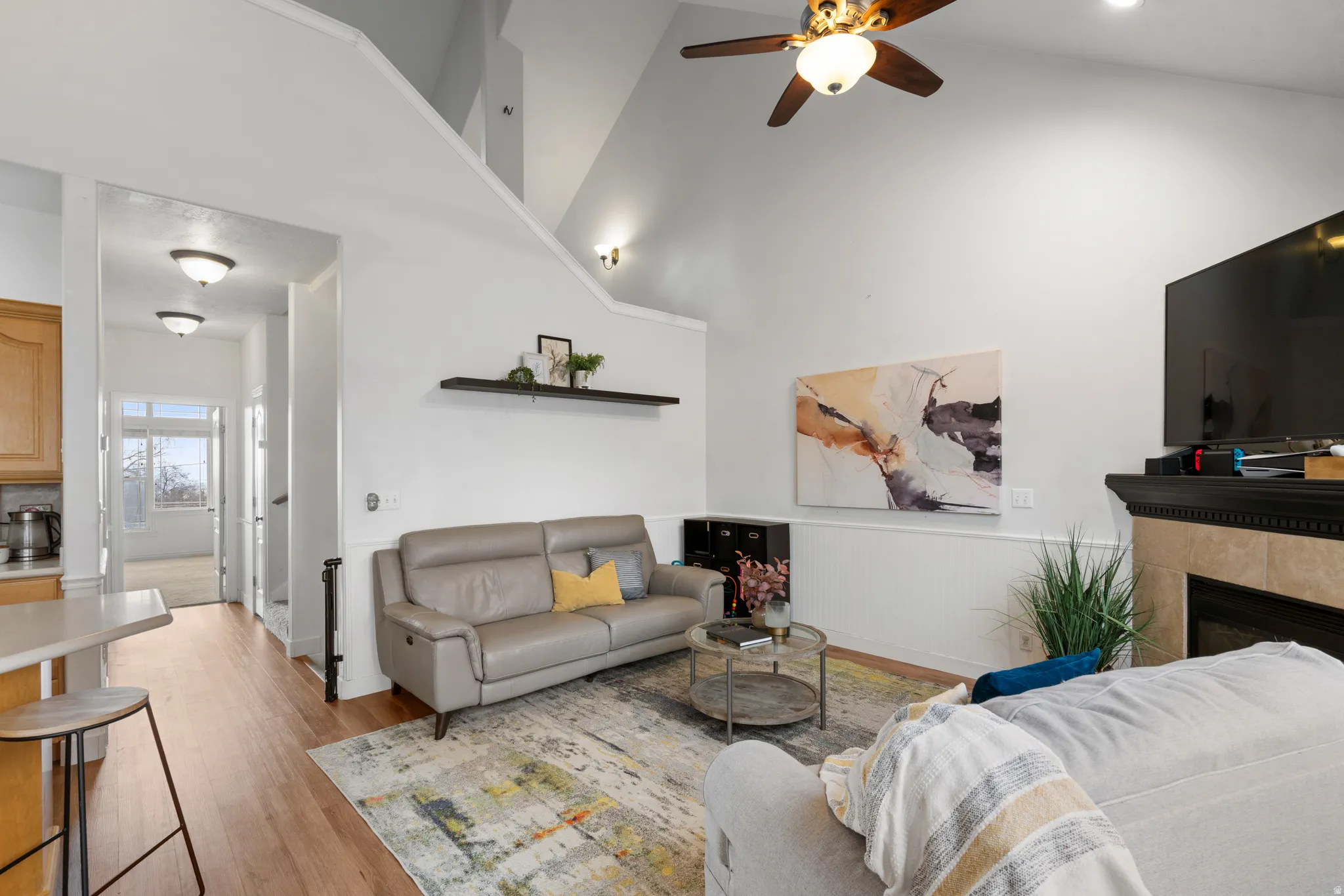 Living room with a fireplace, light wood finished floors, a ceiling fan, a wainscoted wall, and a high ceiling