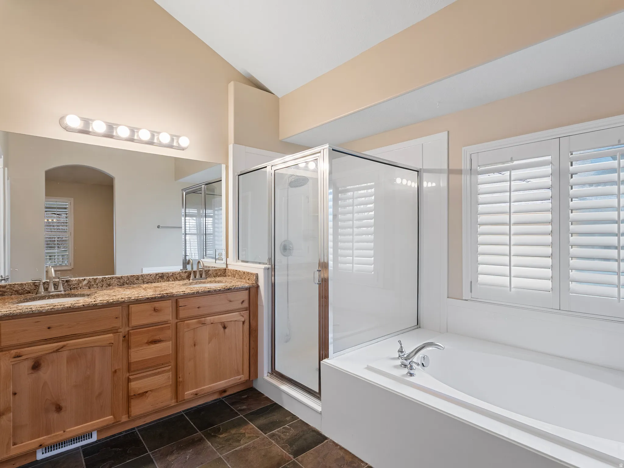 Full bath with double vanity, a shower stall, lofted ceiling, a garden tub, and dark stone finish flooring