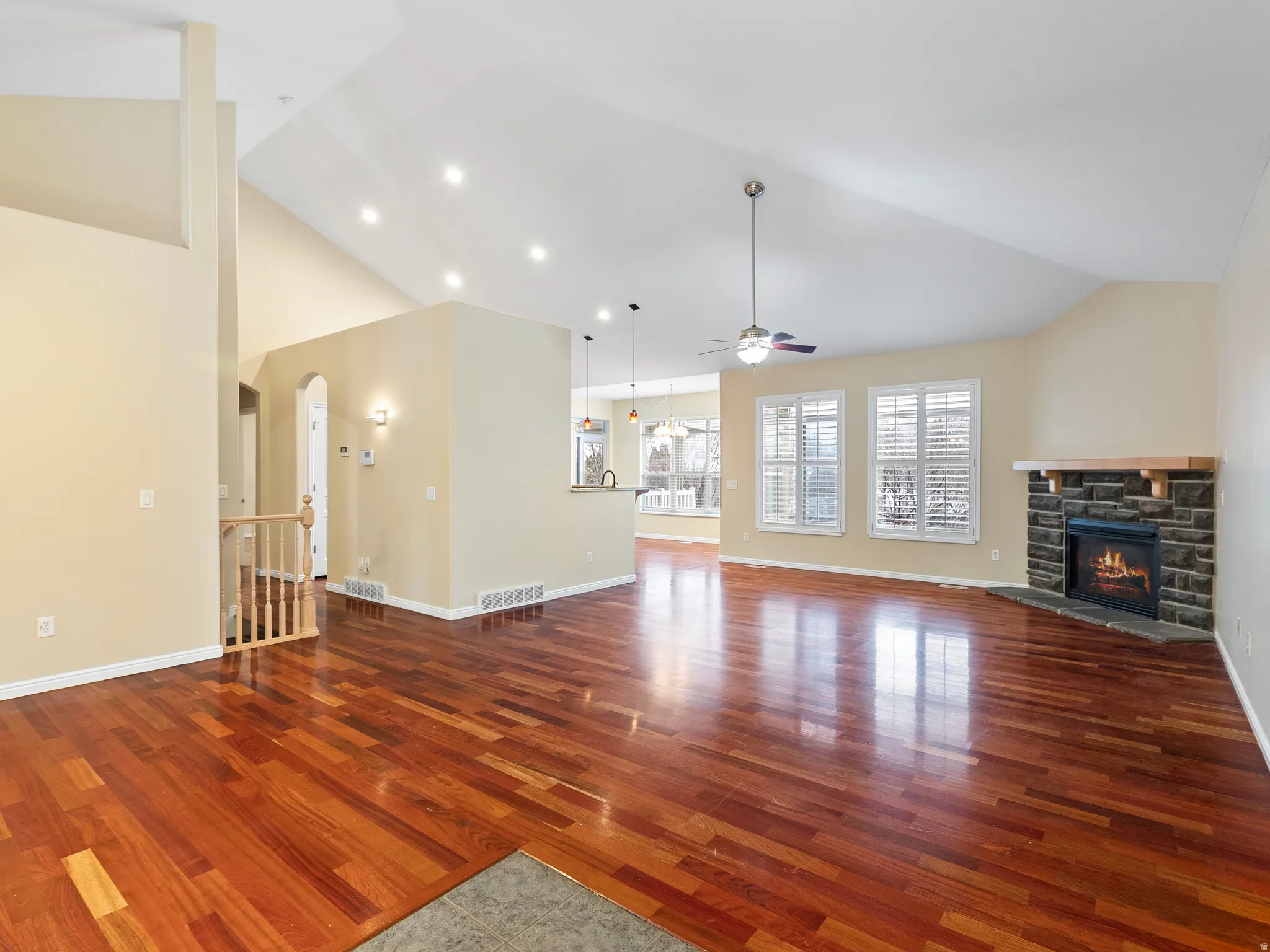 Unfurnished living room featuring dark wood-style floors, vaulted ceiling, a stone fireplace, arched walkways, and a ceiling fan