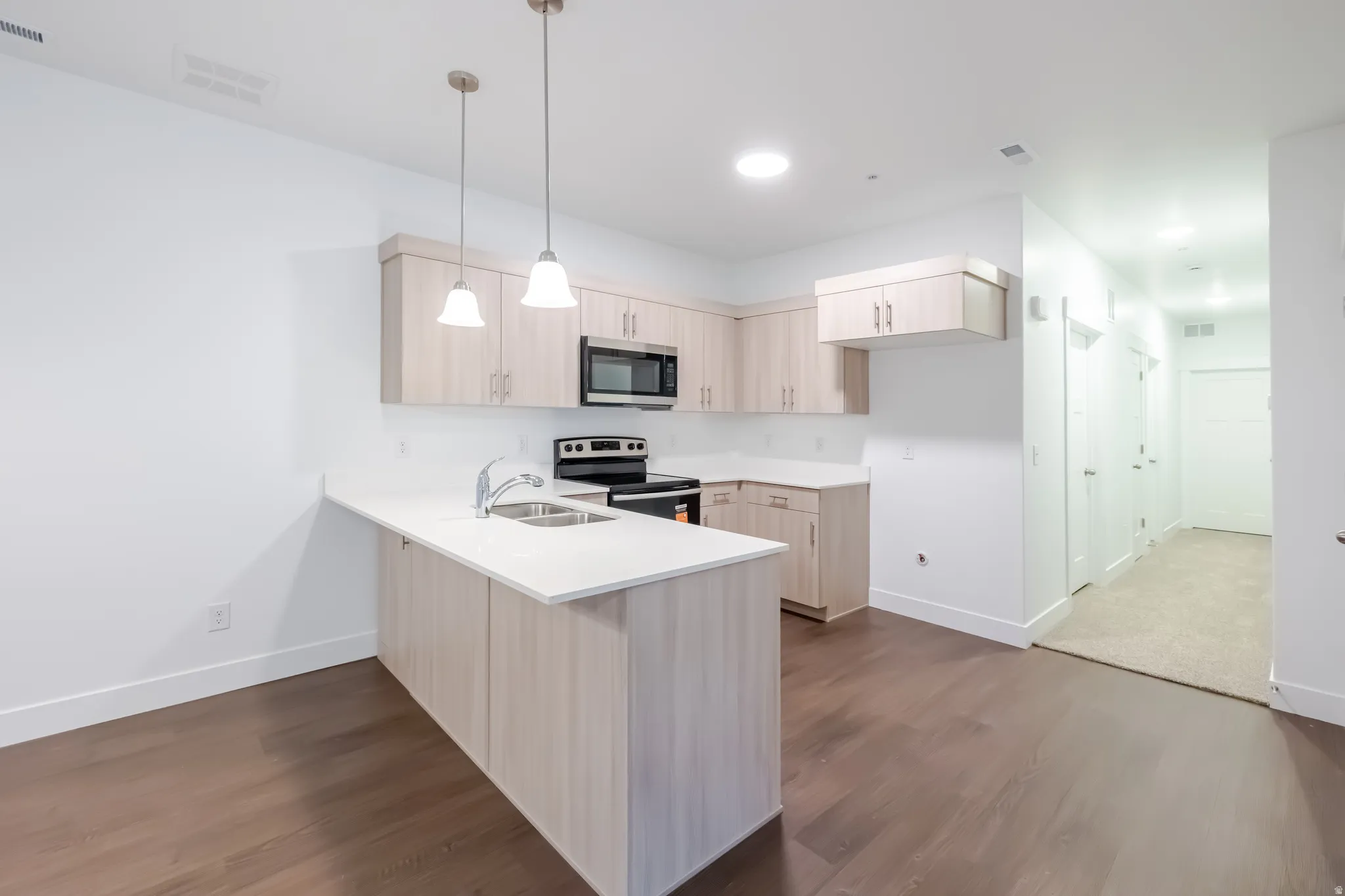 Modern kitchen featuring light wood-finish cabinetry, white quartz countertops, a centered island with seating space, stainless steel appliances including a microwave, pendant lighting, and warm wood-finish flooring throughout the open layout