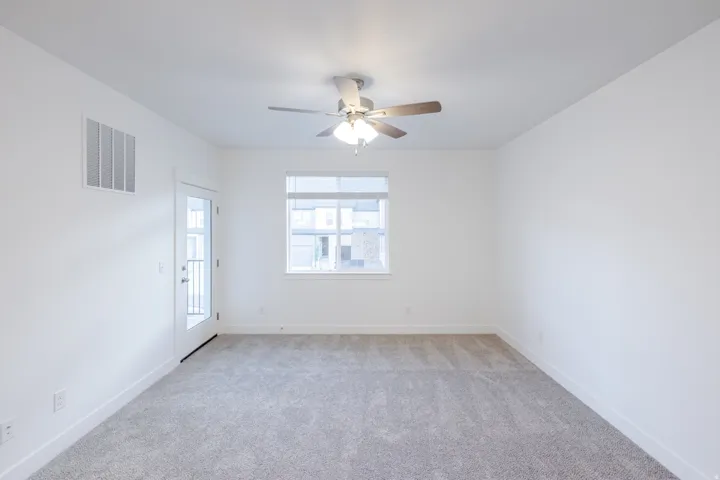 Spacious room with plush gray carpeting, white walls, a ceiling fan with integrated lighting, a window, and a glass-paneled door