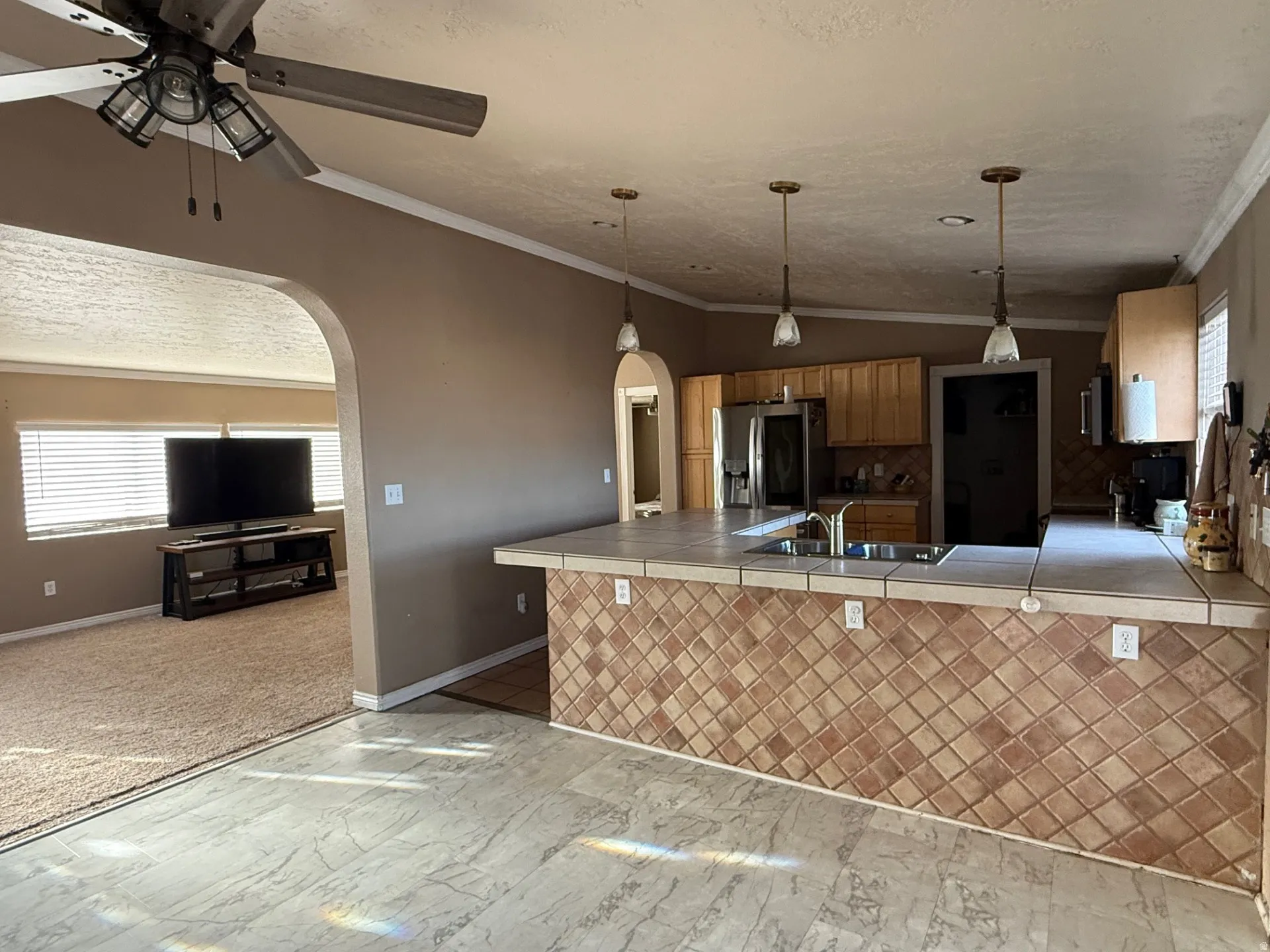 Kitchen with arched walkways, tile countertops, crown molding, stainless steel fridge with ice dispenser, and hanging light fixtures