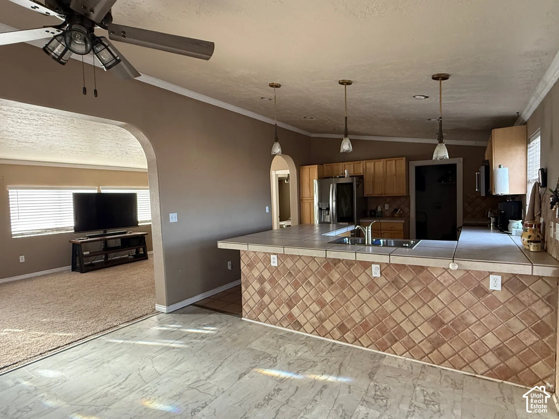 Kitchen with arched walkways, tile countertops, crown molding, stainless steel fridge with ice dispenser, and hanging light fixtures