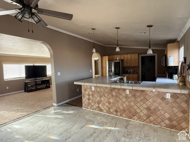 Kitchen with arched walkways, tile countertops, crown molding, stainless steel fridge with ice dispenser, and hanging light fixtures
