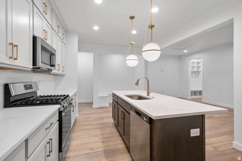 Dual tone kitchen with stainless steel appliances, two tone cabinetry, light wood-type flooring, light stone countertops, and decorative light fixtures