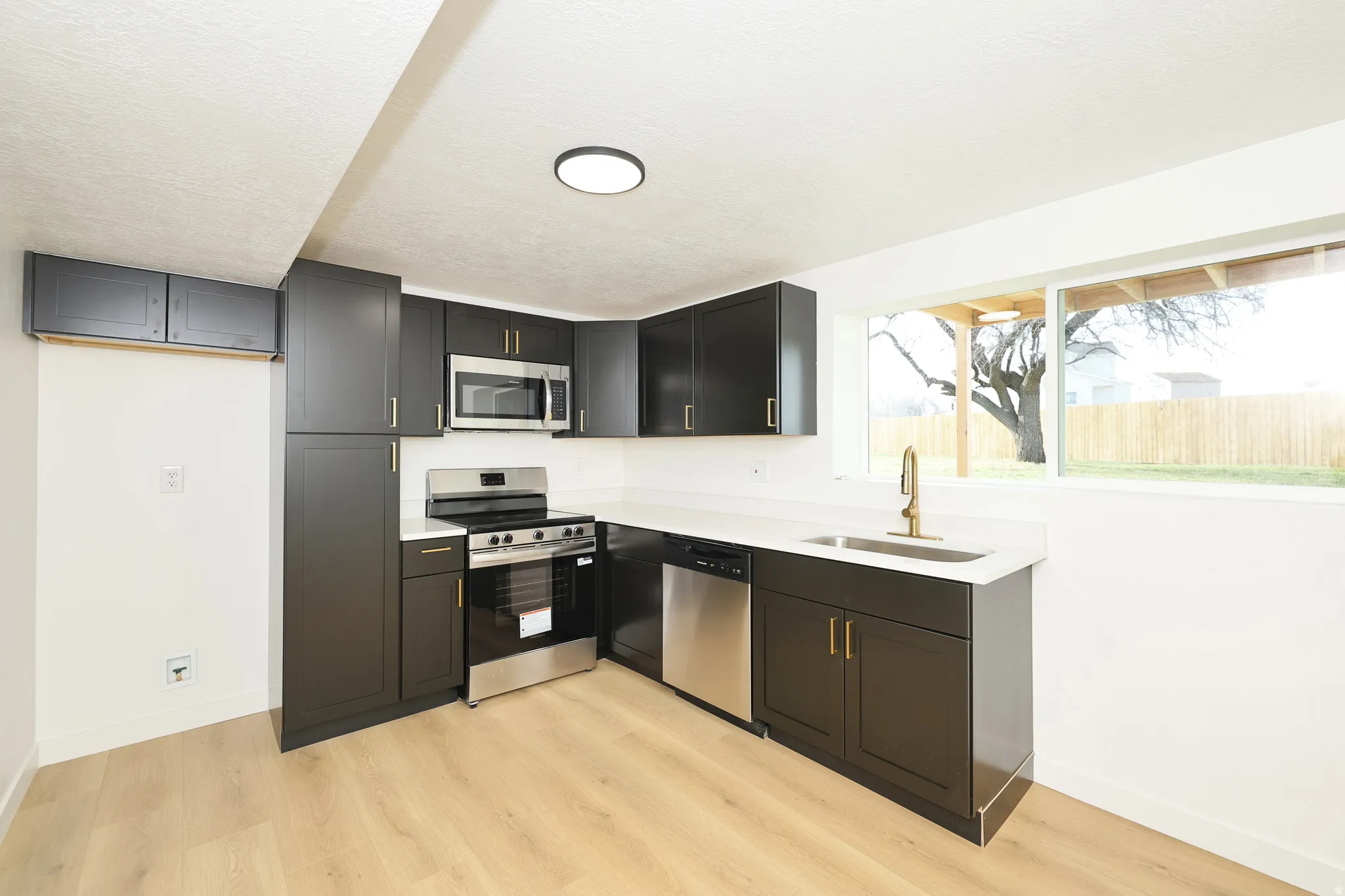 Kitchen with stainless steel appliances, light wood-style flooring, dark cabinetry, light stone countertops, and a textured ceiling