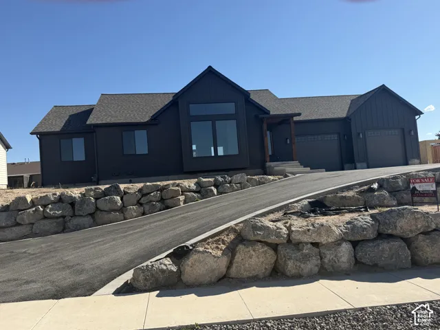 Contemporary exterior featuring dark siding, a multi-pane picture window, an integrated two-car garage, a covered entryway with wood columns, and a paved driveway with stone retaining walls