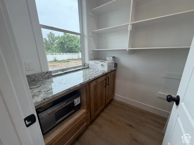 Bright pantry space featuring extensive shelving, granite countertops, wood-finish cabinetry, a large window, and wood-finish flooring