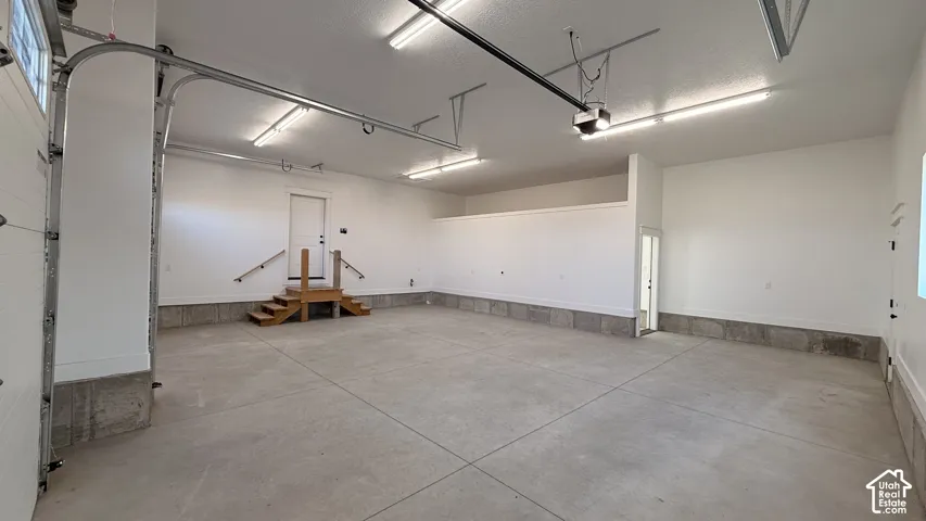 Spacious concrete garage with multiple overhead light fixtures, an exposed garage door track system, and an interior access door with wooden steps and handrails