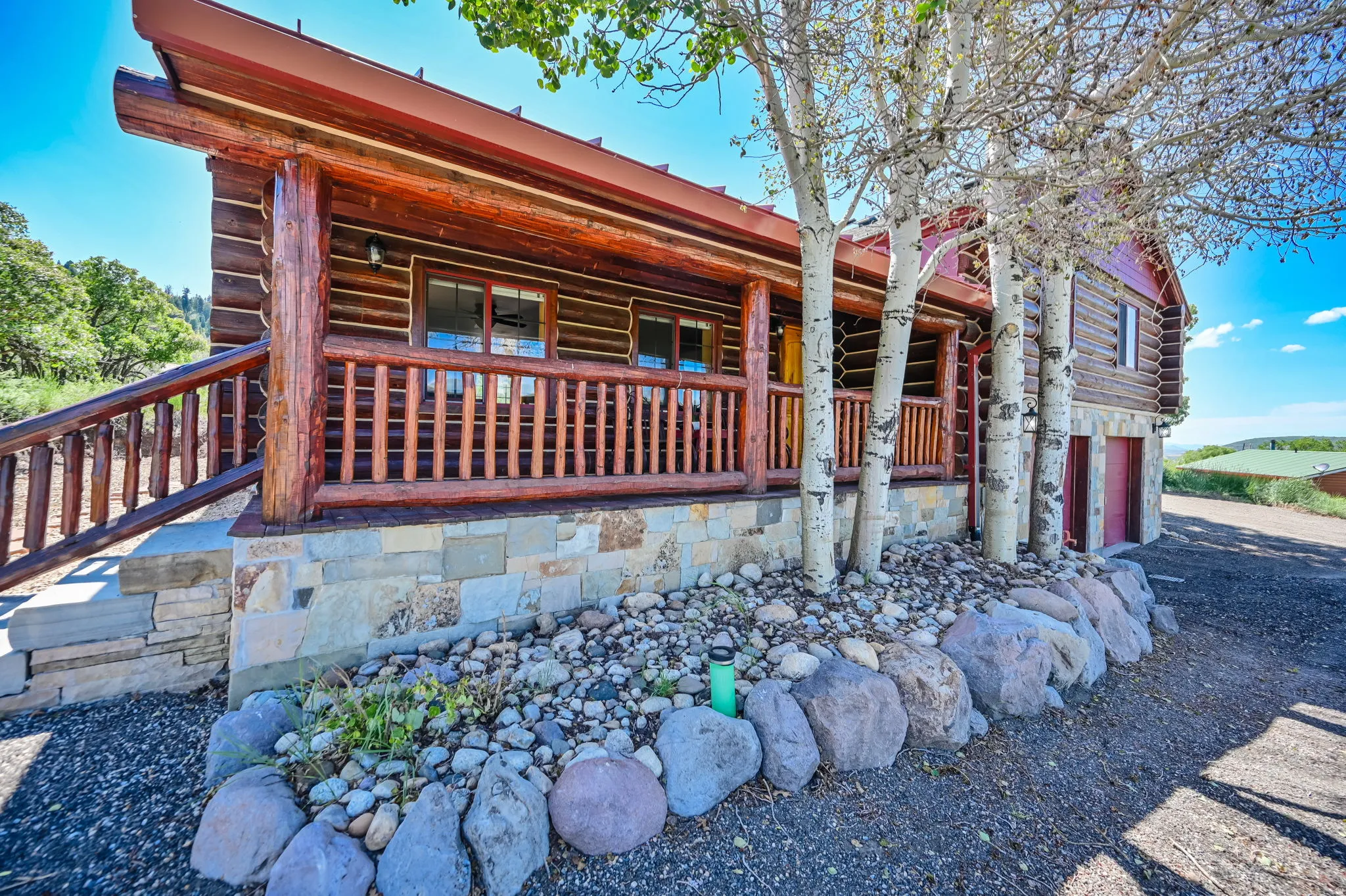 Log cabin exterior featuring a stone foundation, covered front porch with a wood railing, and mature deciduous trees