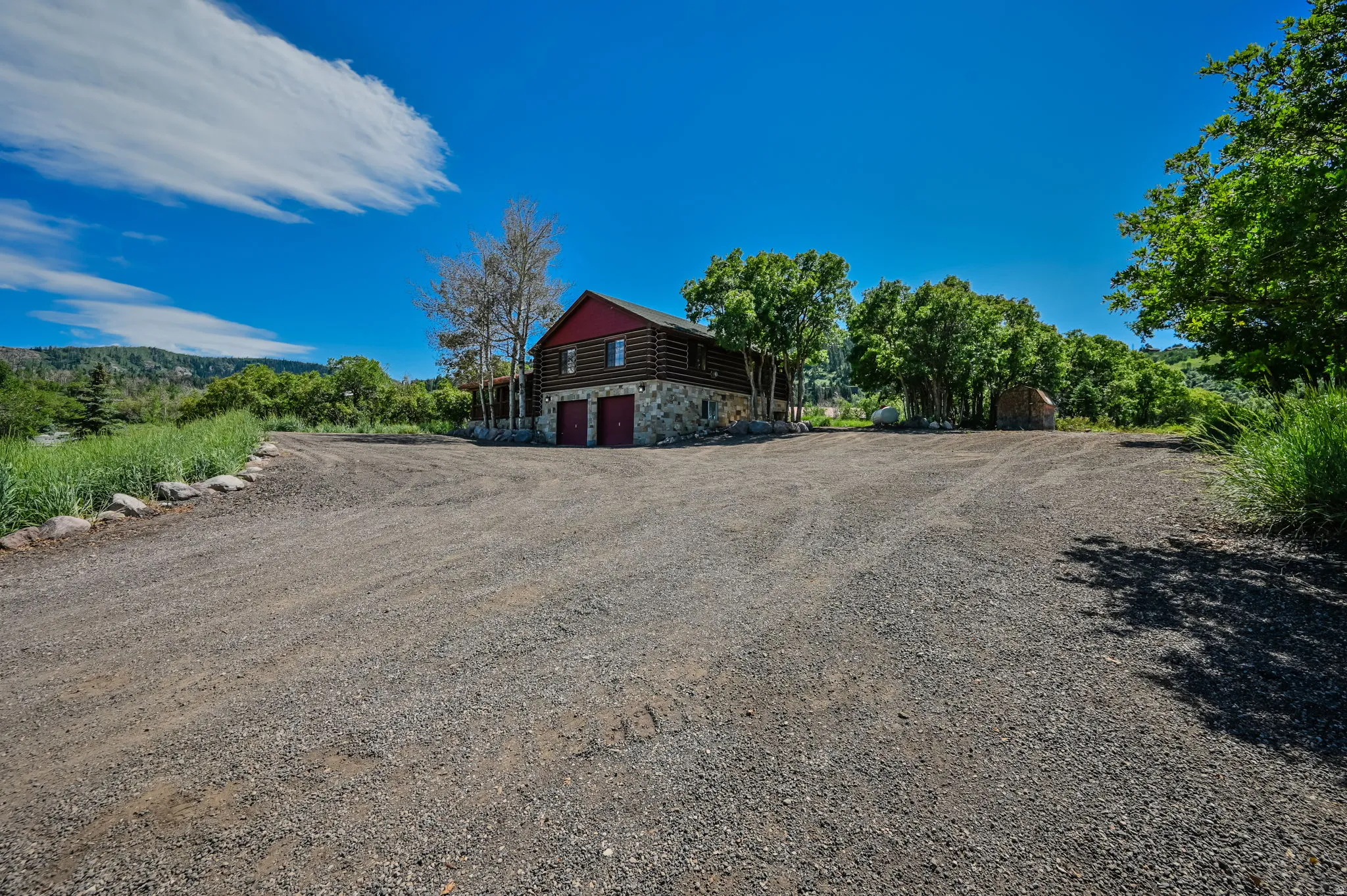 Gravel driveway leading to a two-story log and stone structure with a red roof and two garage doors