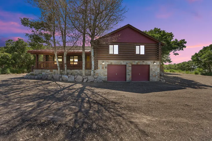 Log cabin exterior featuring a stone foundation, two-car garage with red doors, and a covered front porch with a wooden railing