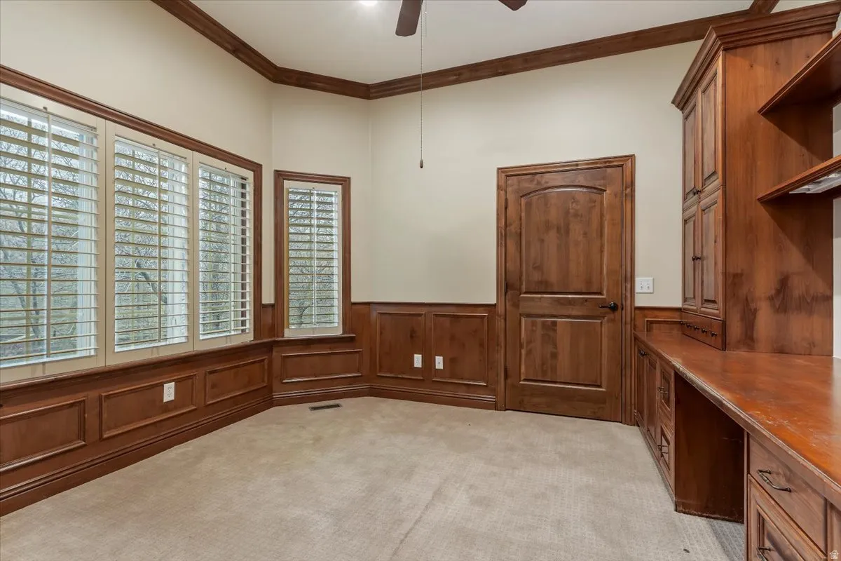 Unfurnished office with ceiling fan, crown molding, light carpet, and wainscoting