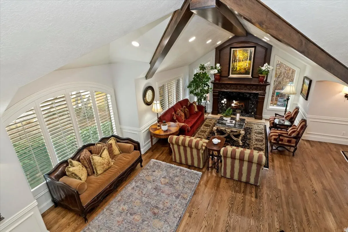 Living room with a warm lit fireplace, wood finished floors, beam ceiling, plenty of natural light, and recessed lighting