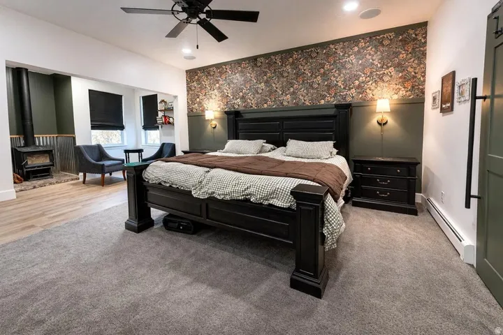 Bedroom with an accent wall, a baseboard radiator, a wood stove, ceiling fan, and light carpet