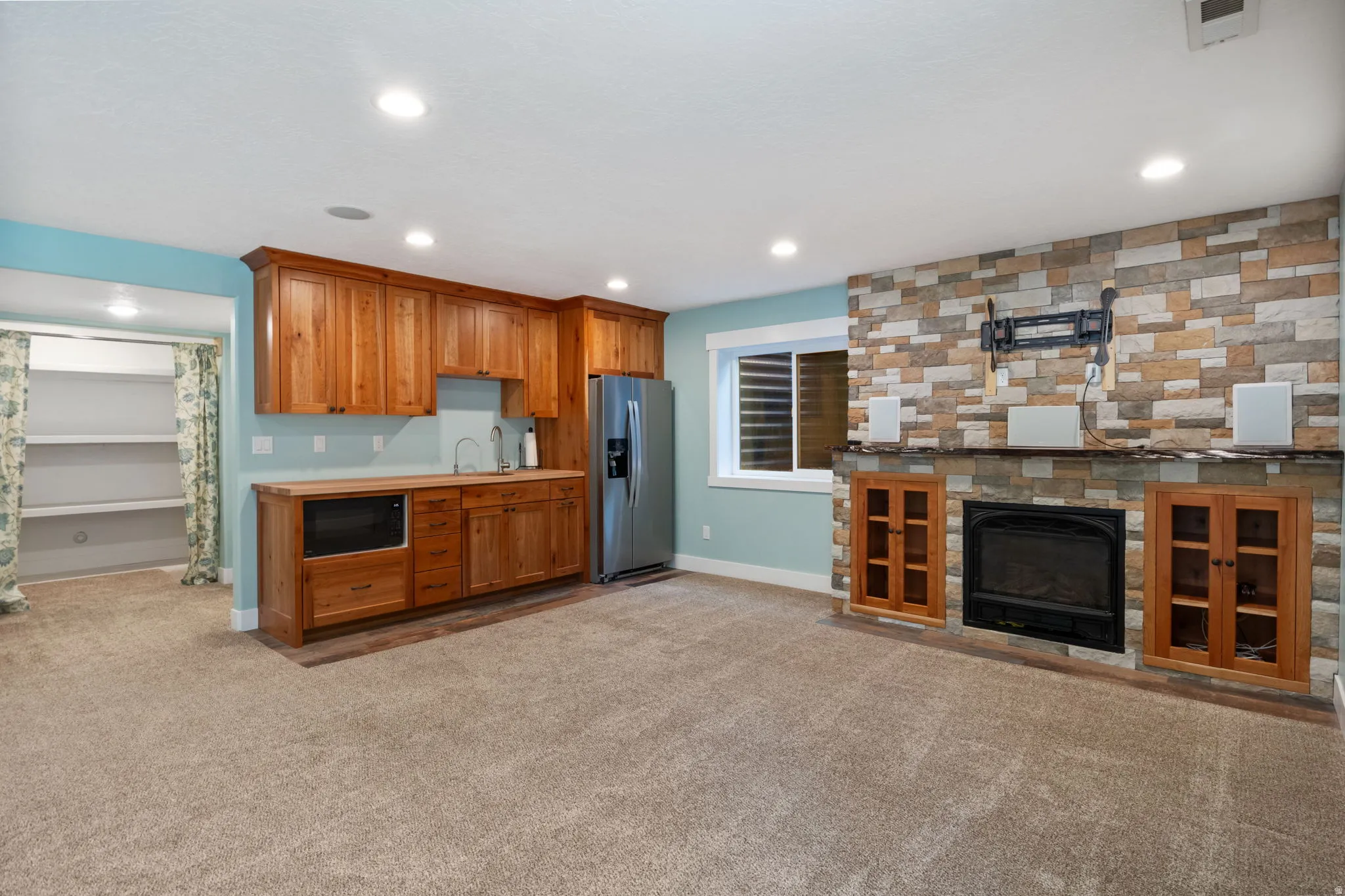 Expansive living area featuring a stone-clad fireplace with built-in cabinetry, kitchenette with wood cabinetry and stainless steel appliances, recessed lighting, and carpet flooring