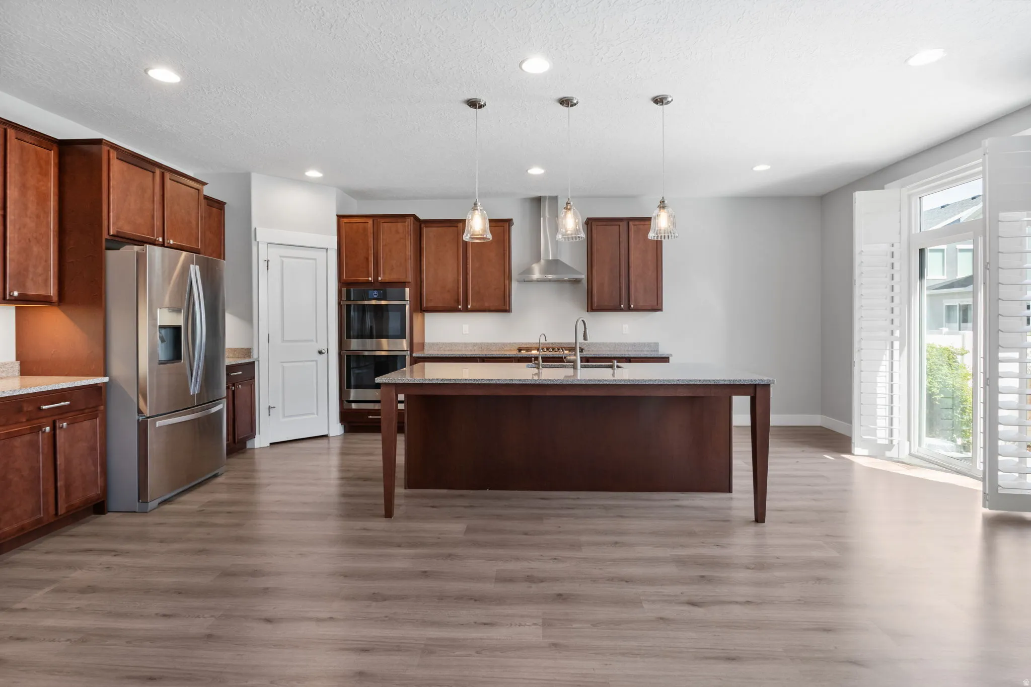 Spacious kitchen featuring rich wood cabinetry, stainless steel appliances including a built-in double oven and refrigerator, a large island with sink, and wood-finish flooring