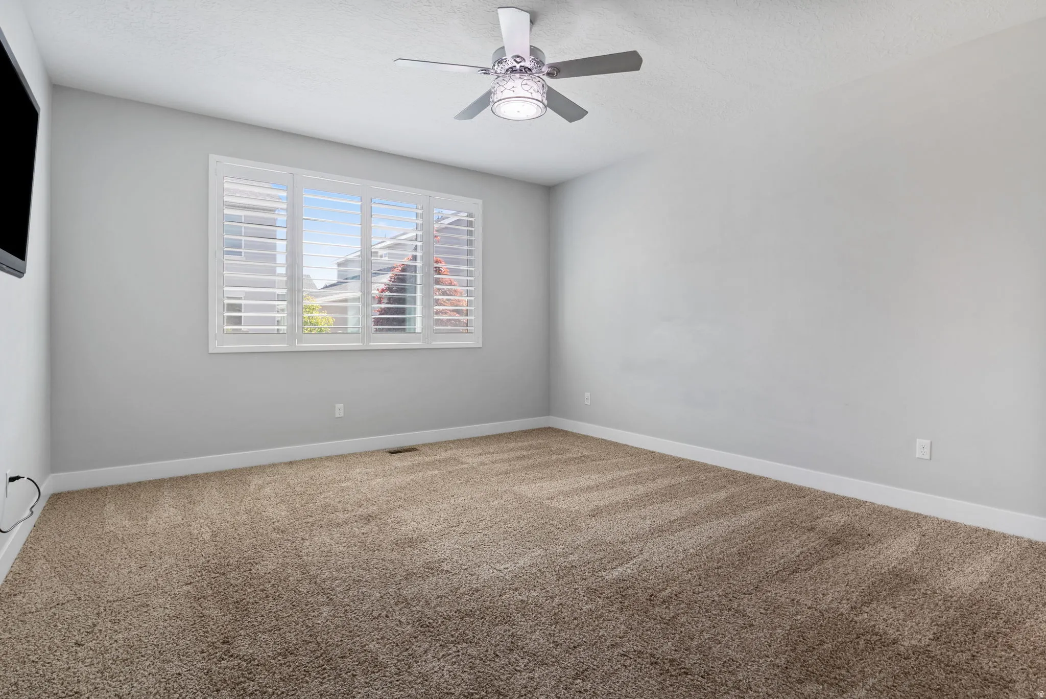 Spacious primary suite featuring light-toned walls, plush carpet flooring, a window with plantation shutters, a modern ceiling fan, and baseboard trim