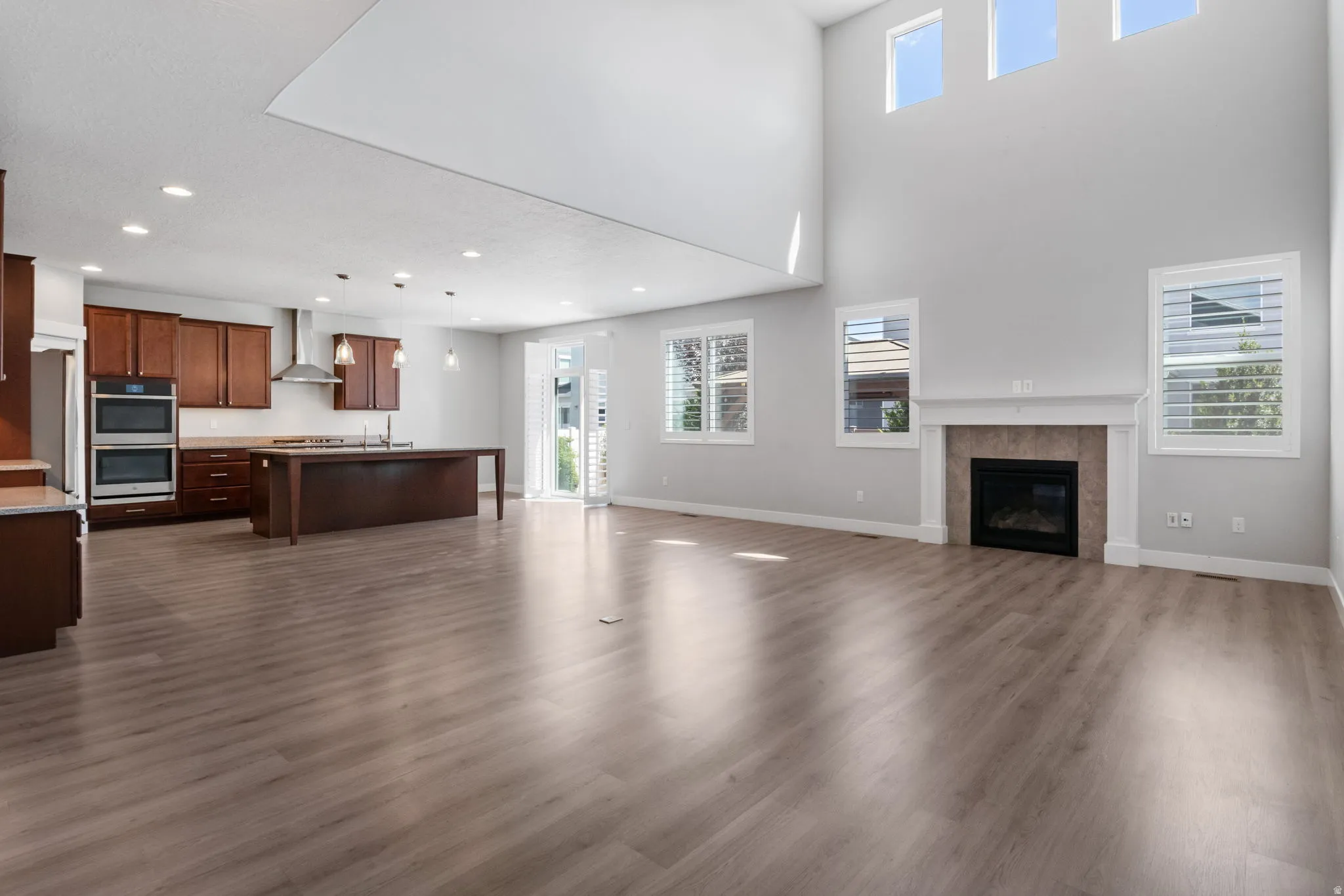 Open-concept living area featuring high ceilings, wood-finish flooring, a contemporary fireplace with a white mantel, and a kitchen with warm-toned cabinetry, stainless steel double ovens, and a large island