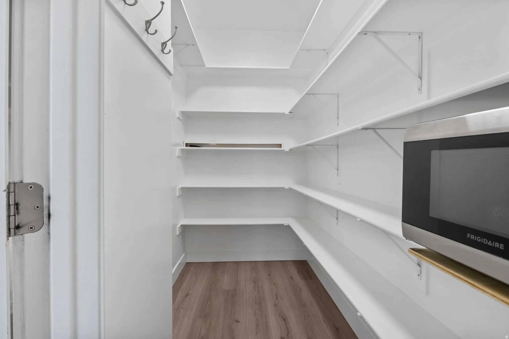 Walk-in pantry featuring white shelving, wood-finish flooring, and a built-in microwave