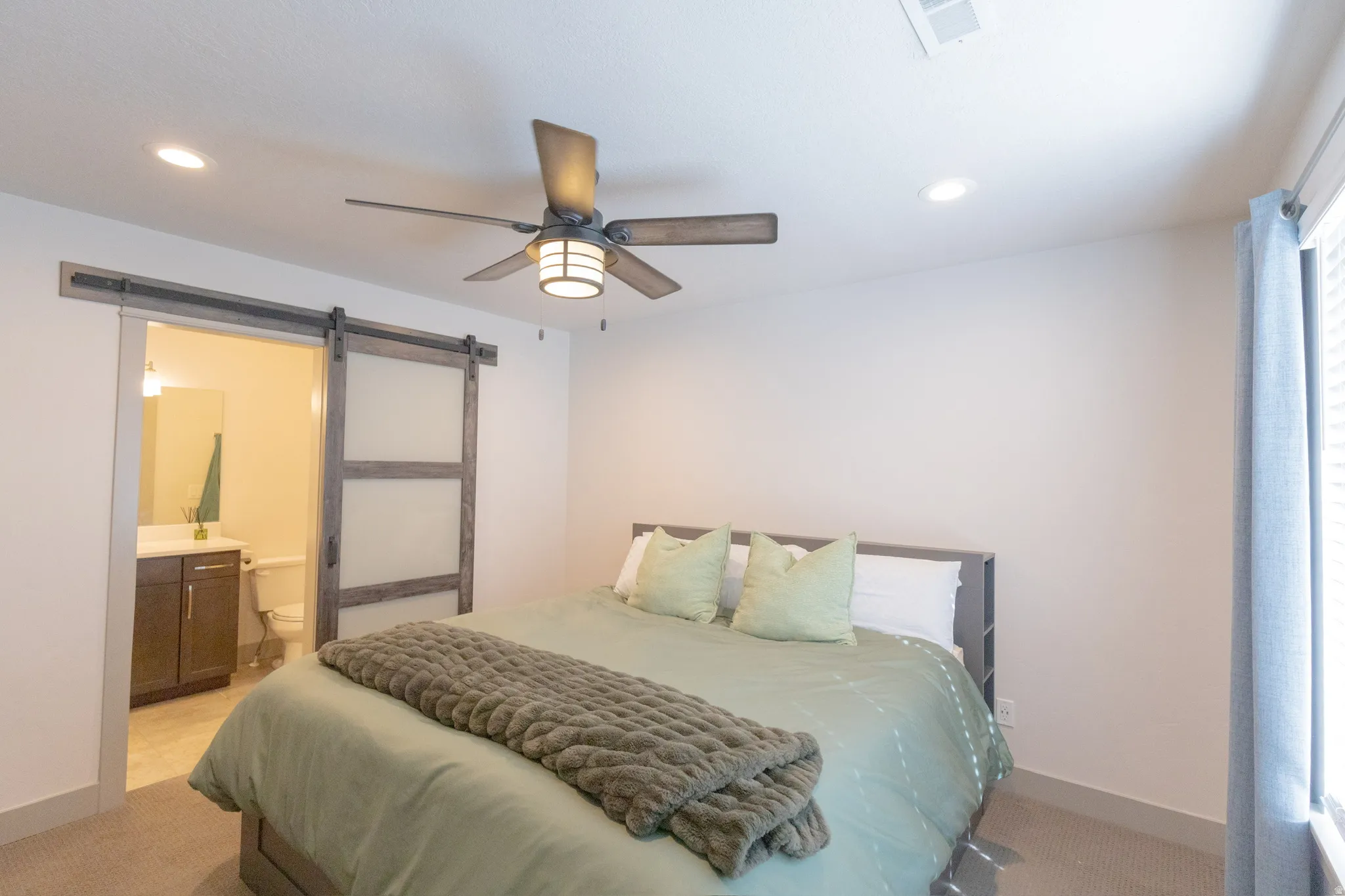 Bedroom with a barn door, light colored carpet, recessed lighting, ceiling fan, and ensuite bathroom