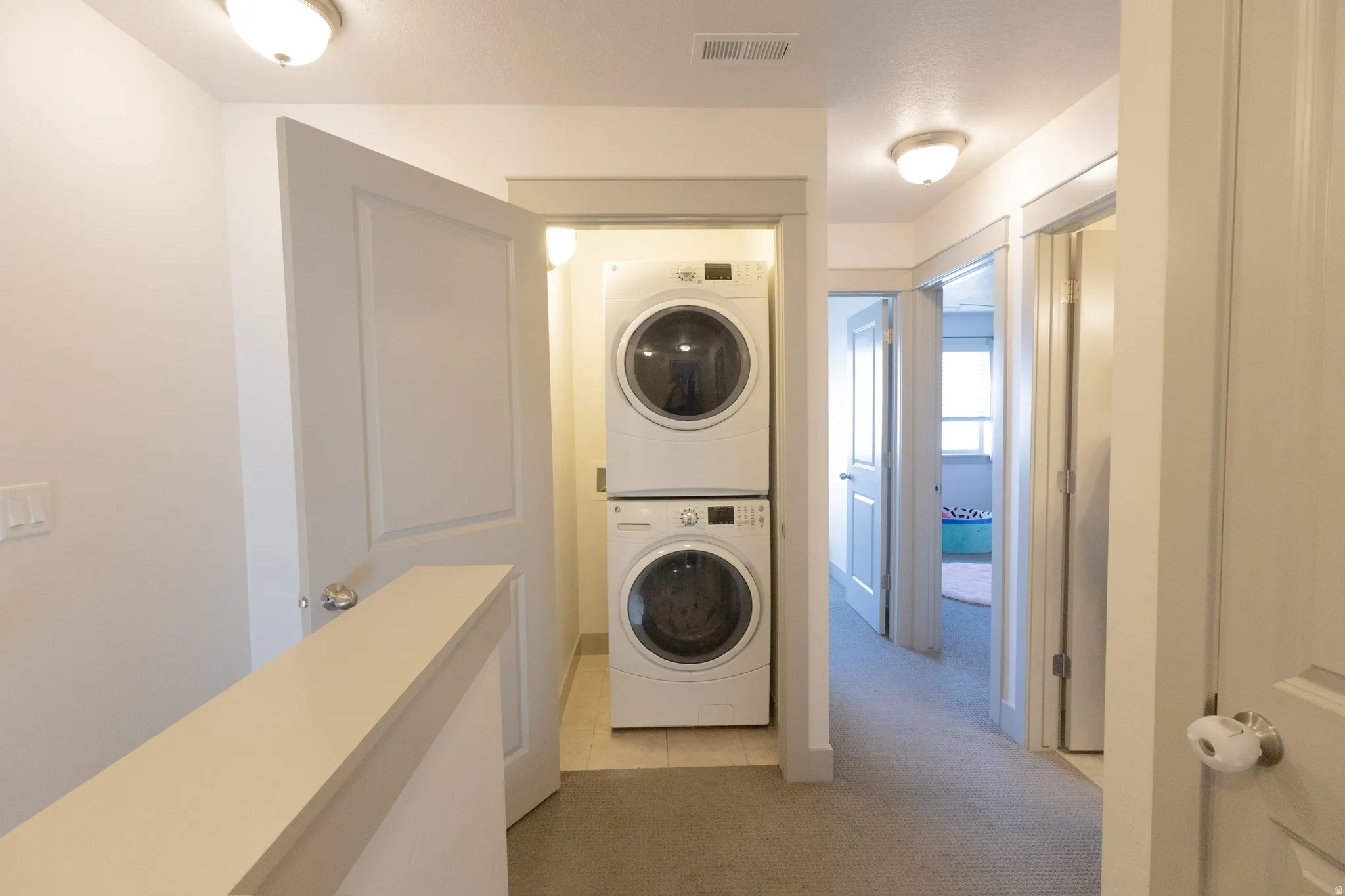 Laundry area featuring light carpet, light tile patterned flooring, and stacked washer / drying machine