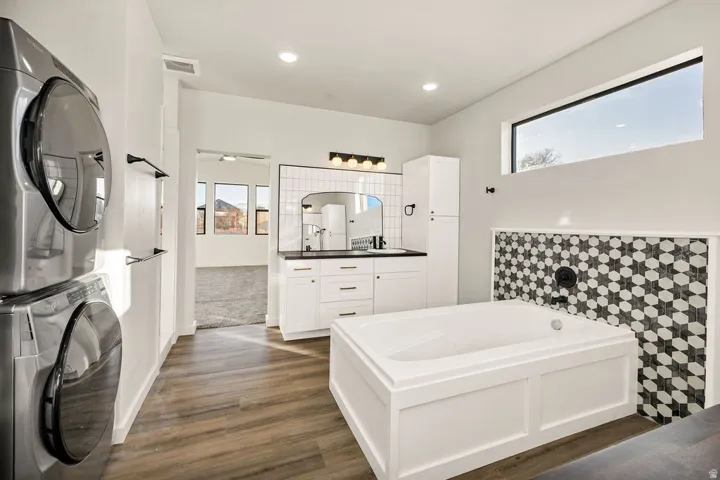 Bathroom with double vanity, a bath, stacked washer / drying machine, dark wood-style flooring, and recessed lighting