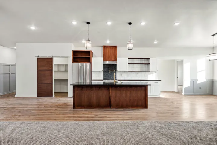 Kitchen featuring open shelves, a barn door, dark countertops, an island with sink, and light wood-type flooring