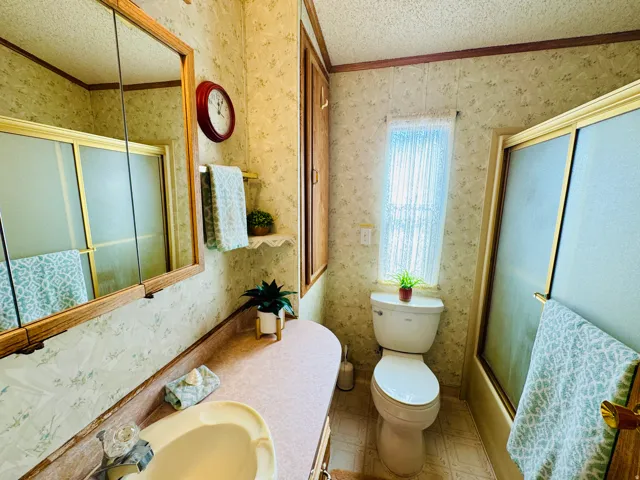 Full bath featuring wallpapered walls, combined bath / shower with glass door, vanity, a textured ceiling, and ornamental molding