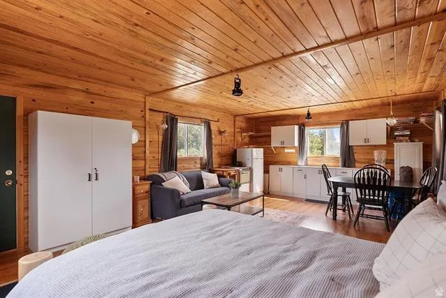 Bedroom with wooden ceiling, multiple windows, light wood-type flooring, freestanding refrigerator, and wooden walls