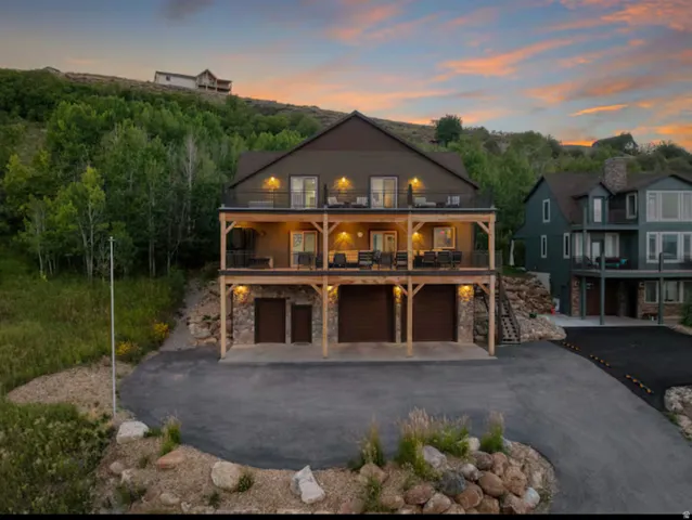 Back of house at dusk with stone siding, a patio area, asphalt driveway, an attached garage, and a balcony