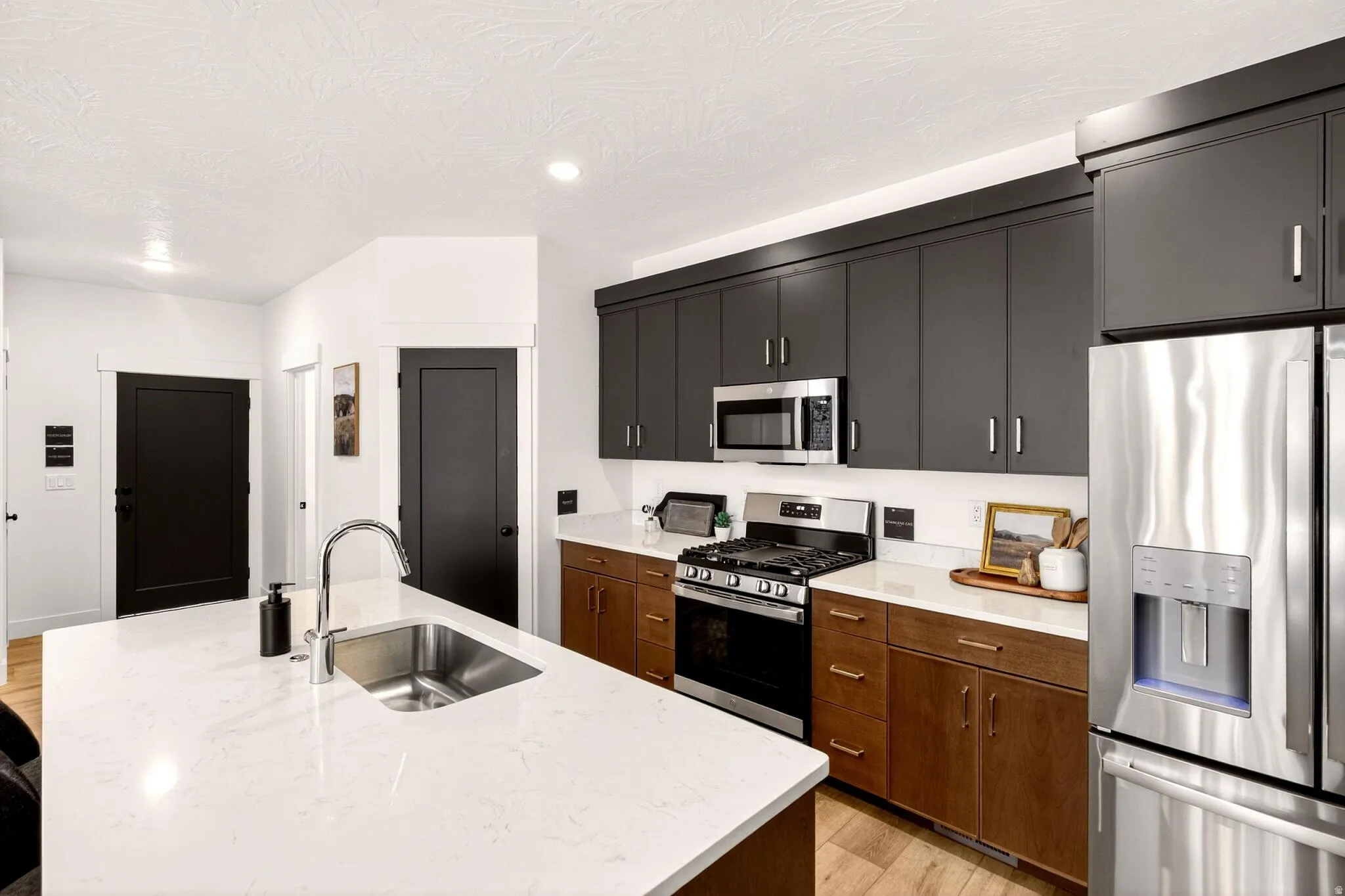 Kitchen with stainless steel appliances, an island with sink, light wood-type flooring, light stone counters, and dark cabinets