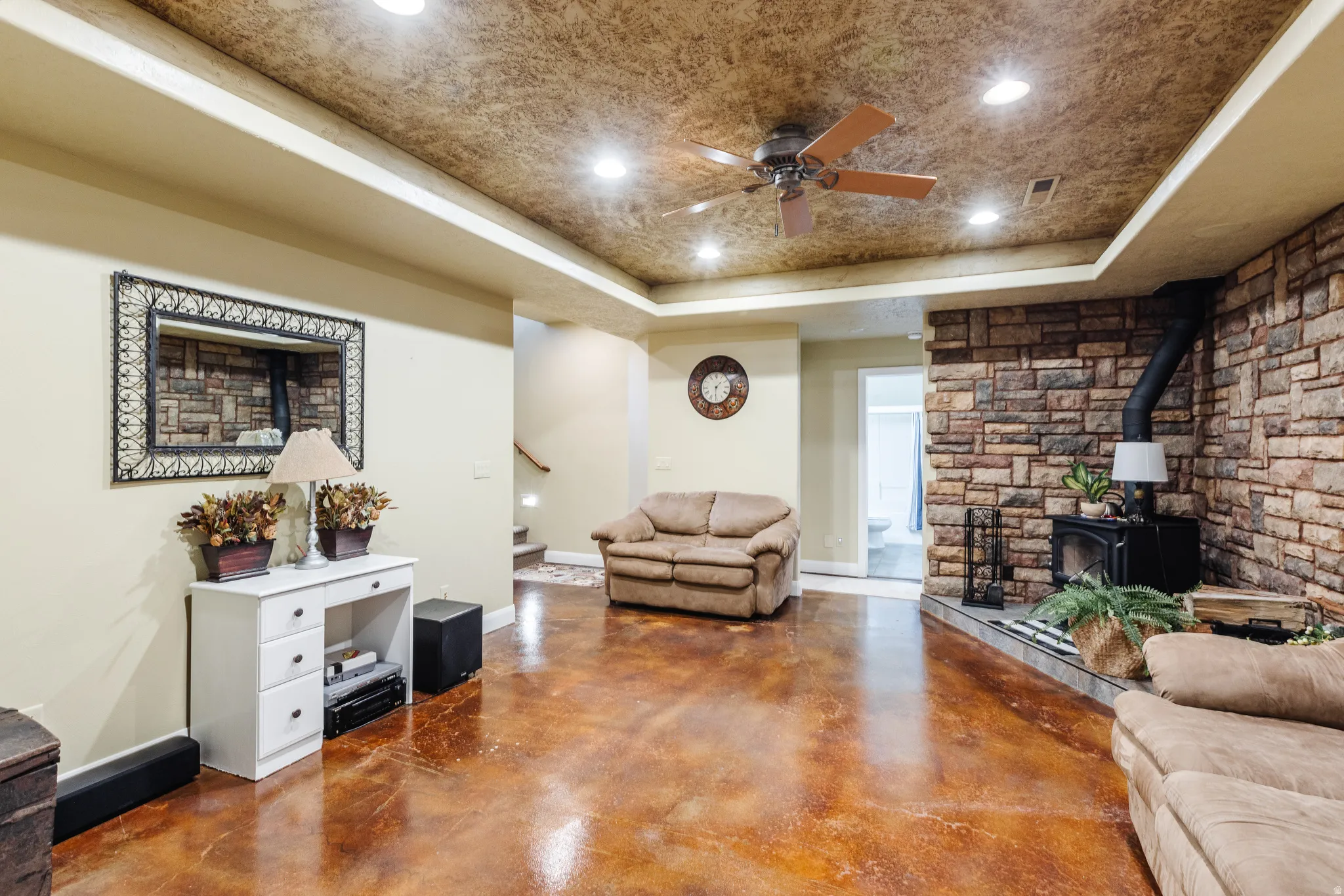 Living room featuring concrete floors, a raised ceiling, a wood stove, ceiling fan, and recessed lighting