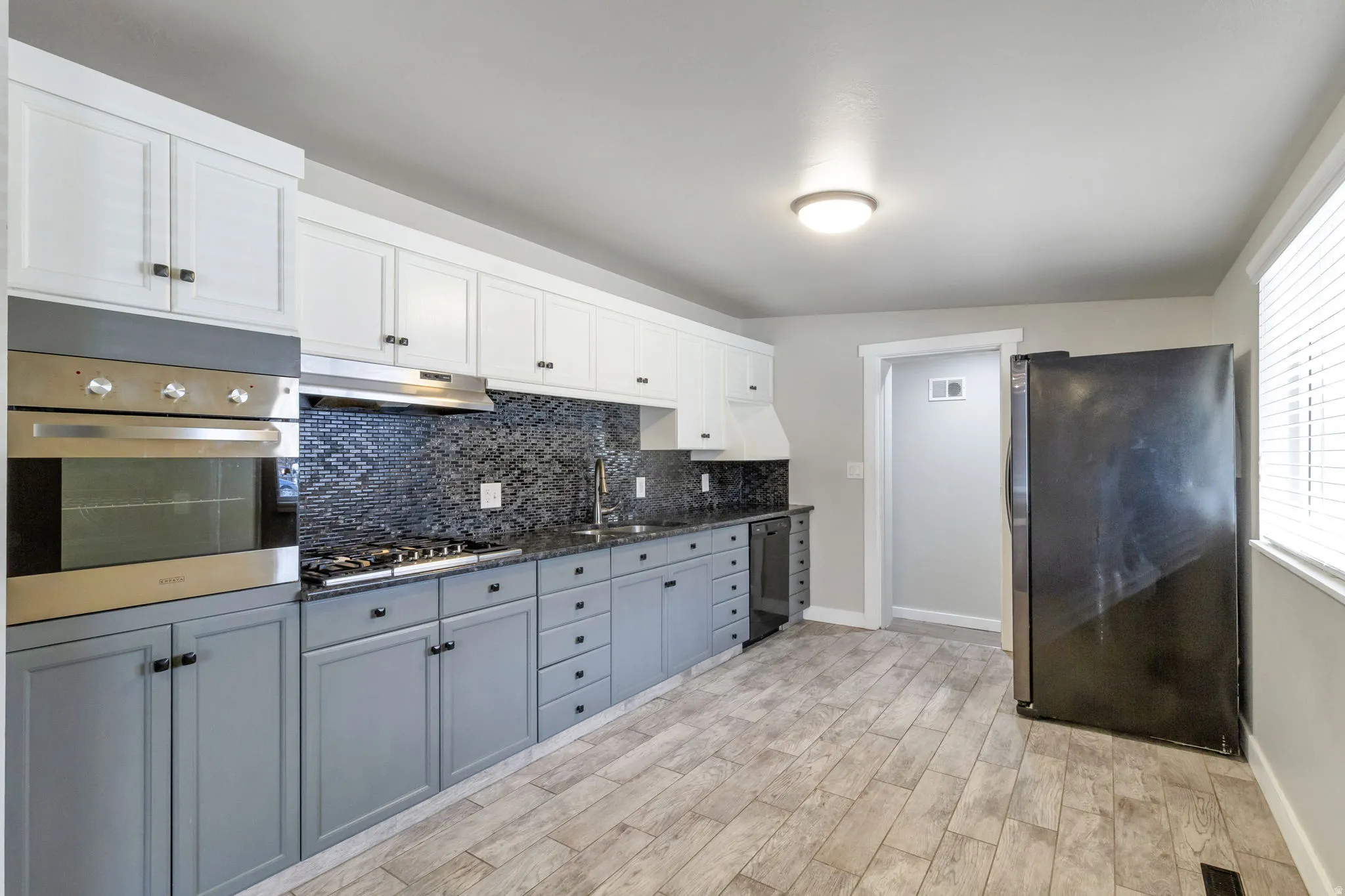 Two tone kitchen with stainless steel appliances, tasteful backsplash, two tone cabinets, light wood-style flooring, and dark stone countertops