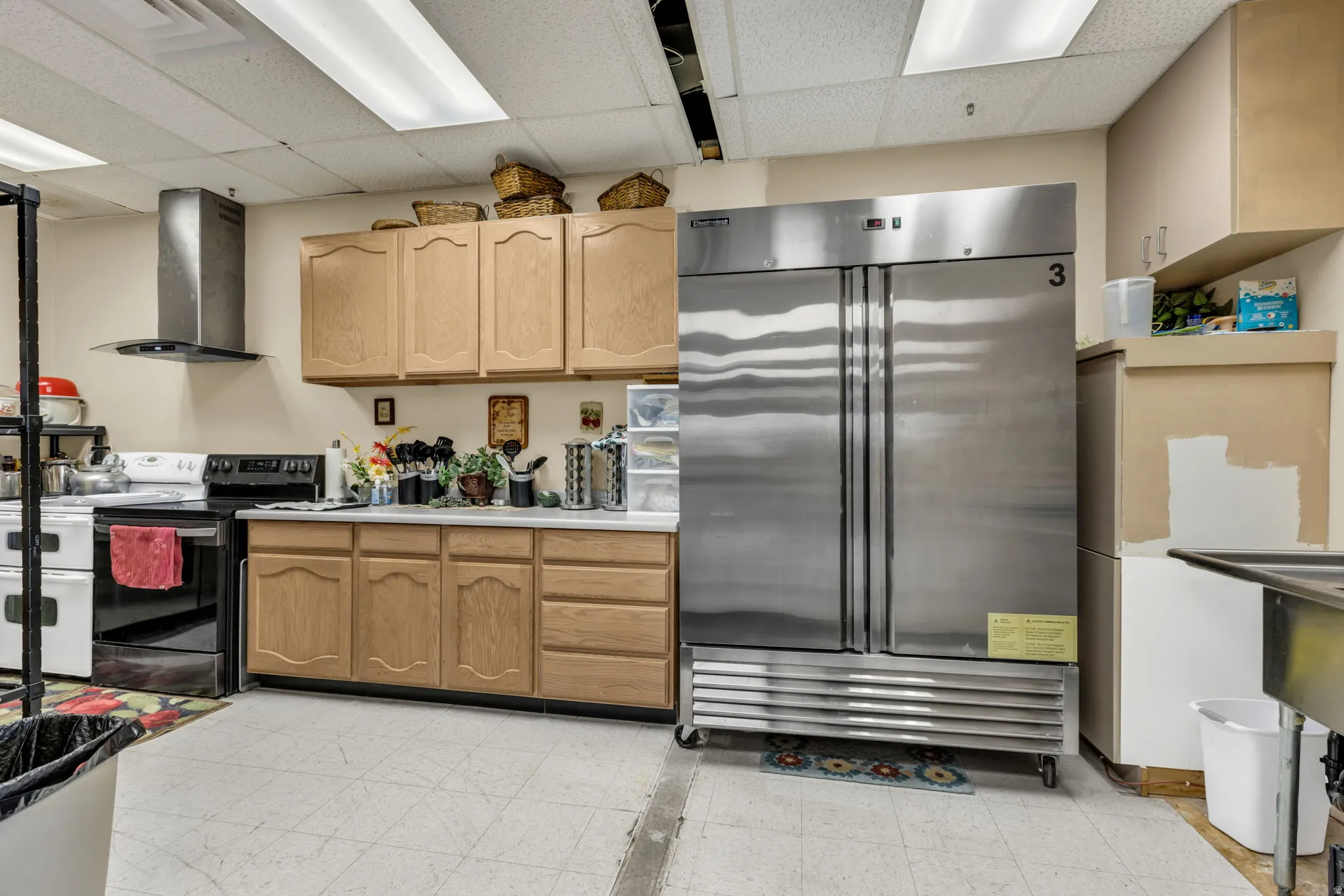 Kitchen with a paneled ceiling, stainless steel refrigerator, electric stove, light countertops, and light wood finish cabinetry