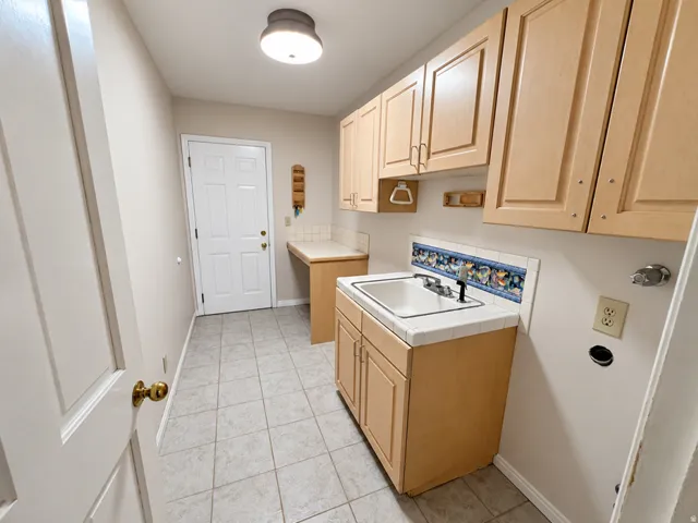 Full-size utility sink with decorative tile backsplash, ample upper and lower cabinetry, tile flooring, and integrated countertop space