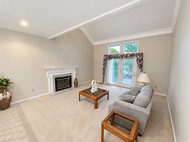 Vaulted ceiling with exposed beams, fireplace with a white mantel, glass-paneled double doors, and light neutral carpeting