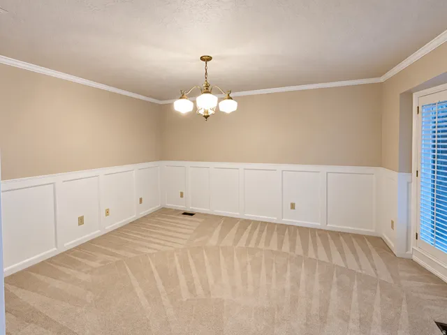 Neutral-toned carpet flooring, crown molding, wall paneling, and a brass chandelier with three glass shades