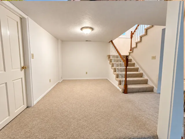Light-colored carpeted flooring, white walls, and a staircase with a wooden handrail and white balusters