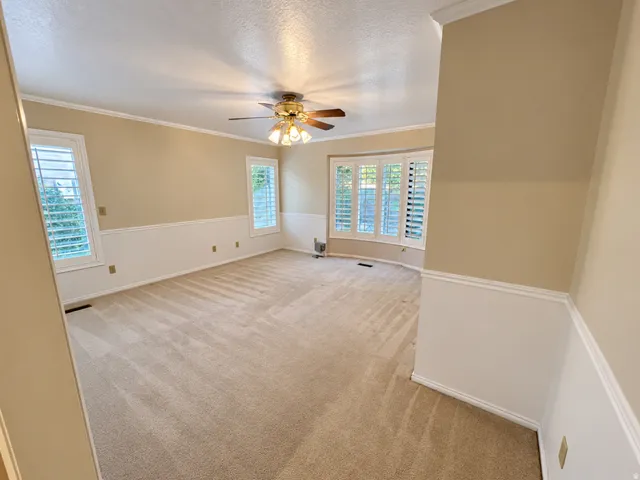 Bay window with plantation shutters, ceiling fan with light fixture, chair rail molding, crown molding, and carpeted flooring