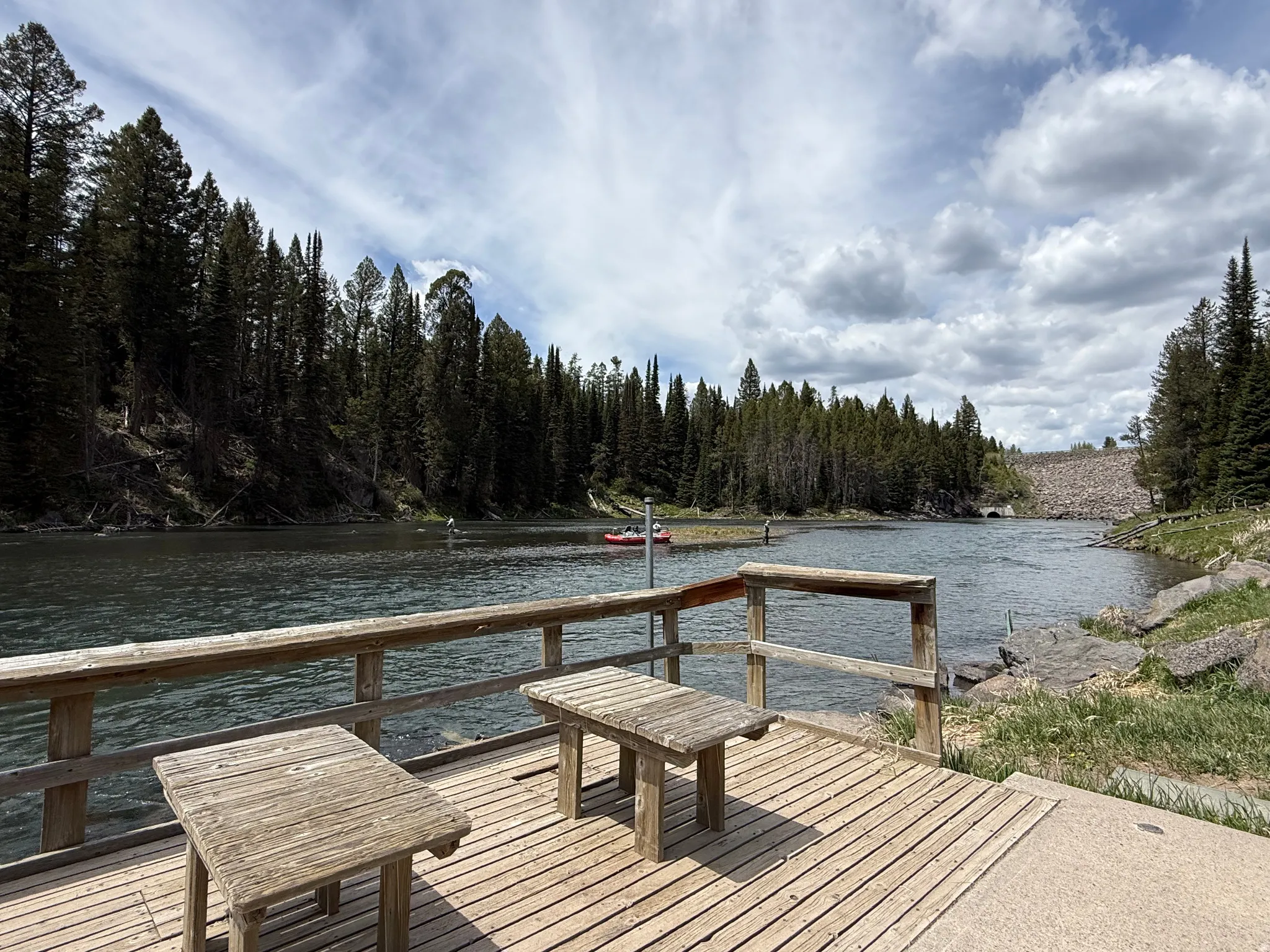 Wood-plank dock with rustic wooden tables, overlooking a river bordered by dense evergreen forests and a distant dam structure
