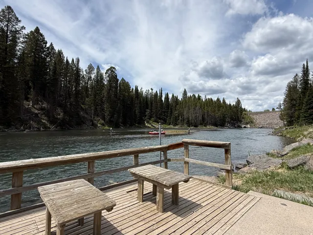 Wood-plank dock with rustic wooden tables, overlooking a river bordered by dense evergreen forests and a distant dam structure