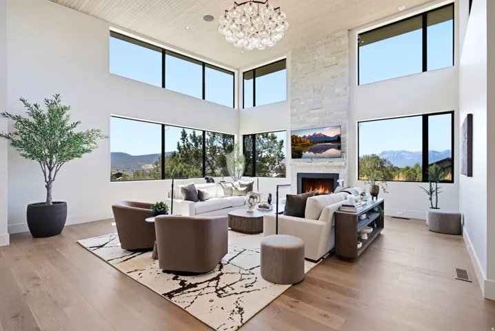 Double-height ceilings, large windows with black framing, wood-finish flooring, a stone-clad fireplace, and a contemporary chandelier