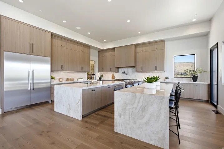 Well-appointed kitchen featuring light wood-finish cabinetry, two waterfall-edge islands with stone countertops, and integrated stainless steel appliances
